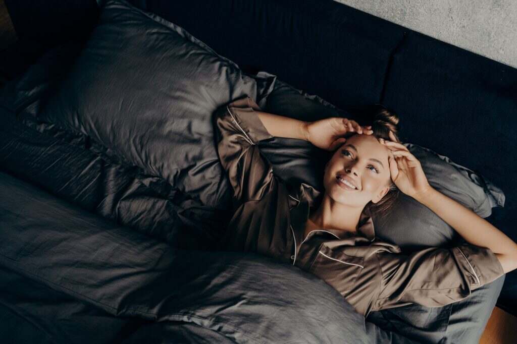 Beautiful young girl in satin comfy pajama waking up while lying in bed at home