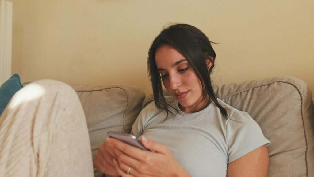 Young woman using mobile phone and changing channels from remote control while sitting