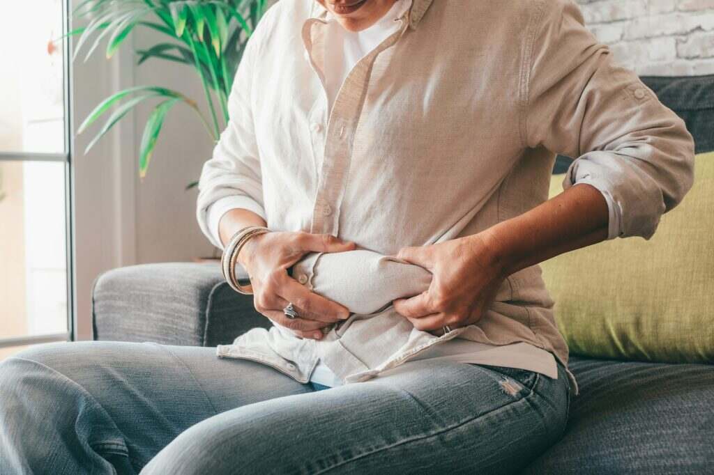Midsection of woman hands holding her belly fat sitting on sofa at home.