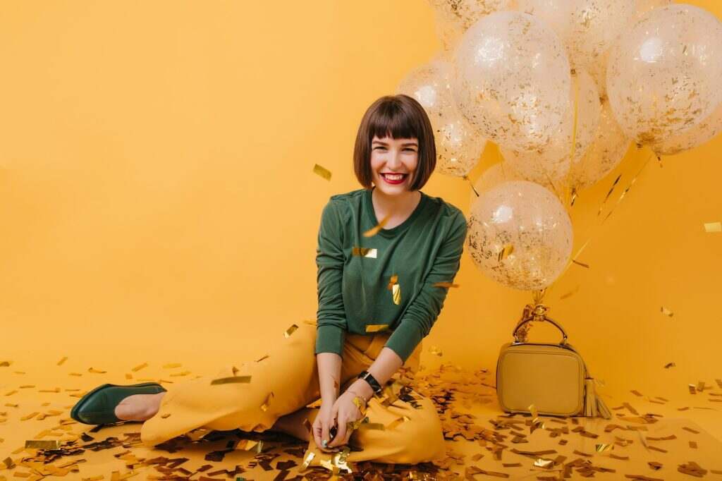 Blissful white girl laughing and looking at golden confetti. Indoor photo of cheerful brunette mode