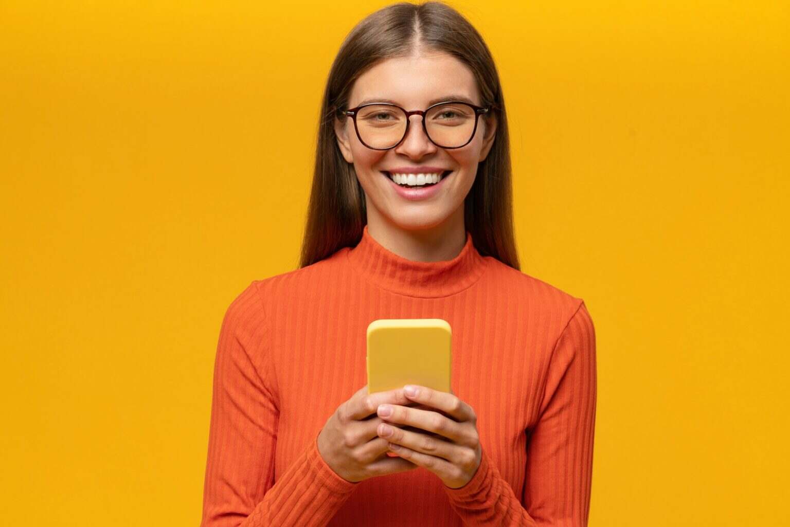 Woman in glasses posing on yellow background using phone laughing at memes in social media