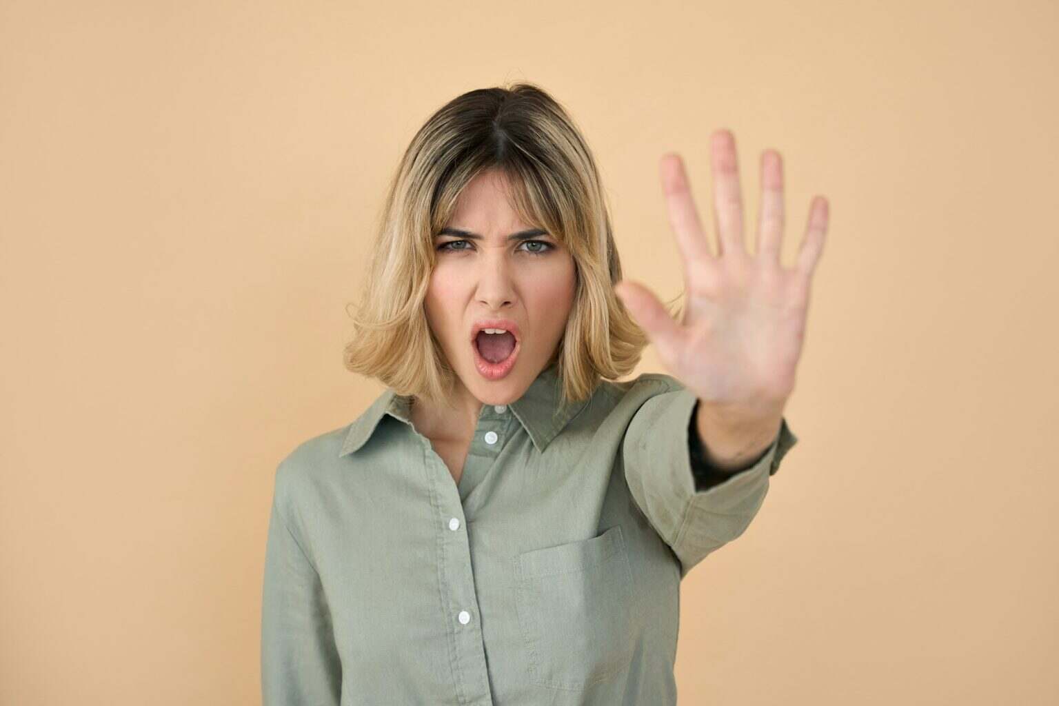Angry blonde young woman screaming showing stop hand gesture isolated on beige.