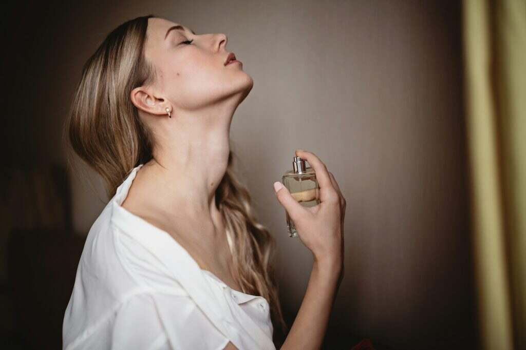 Comment reconnaître un vrai parfum d’un faux ? Les astuces infaillibles Beautiful Caucasian woman with blonde hair spraying luxury perfume from bottle on neck at home