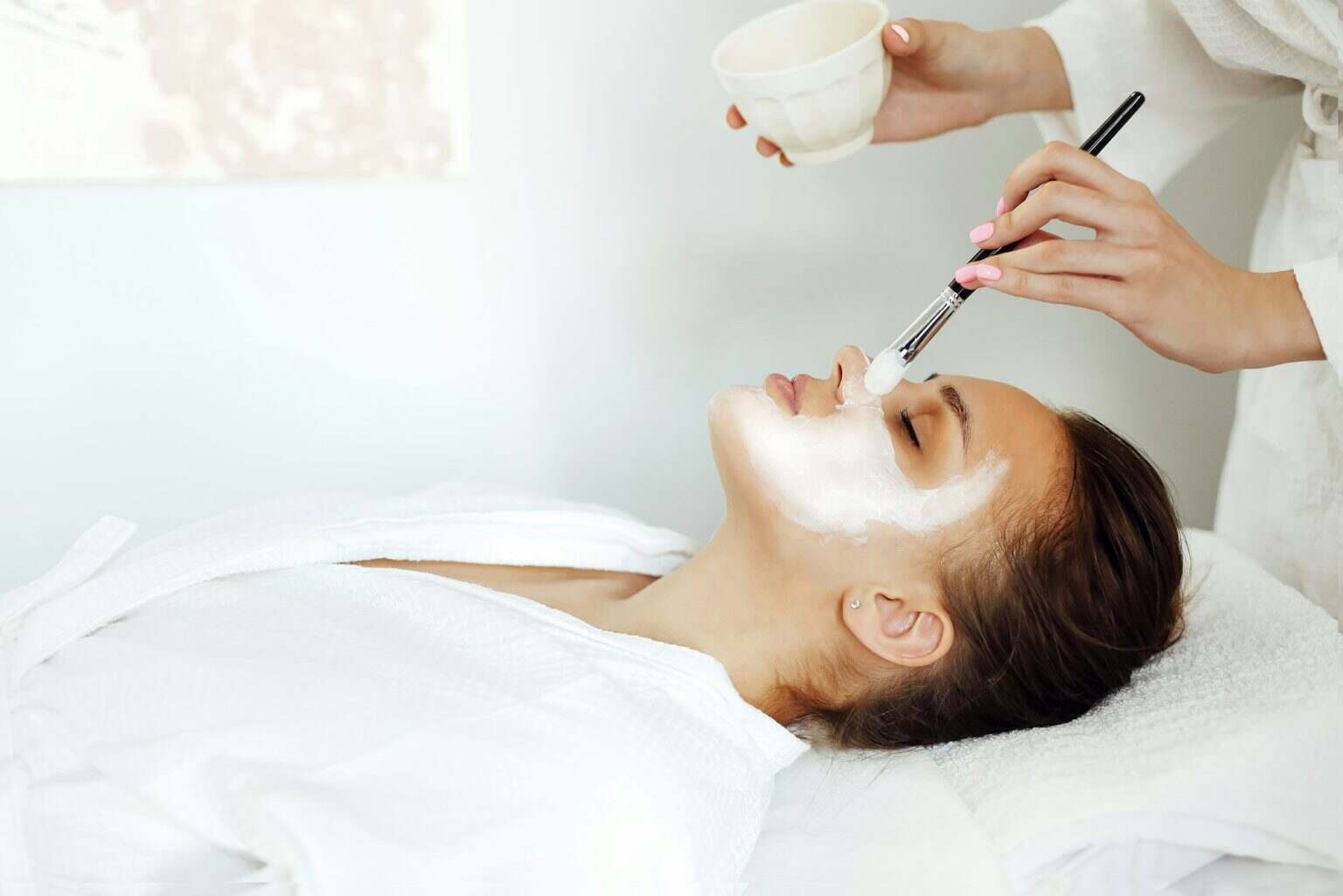 Crop cosmetologist applying facial mask on client