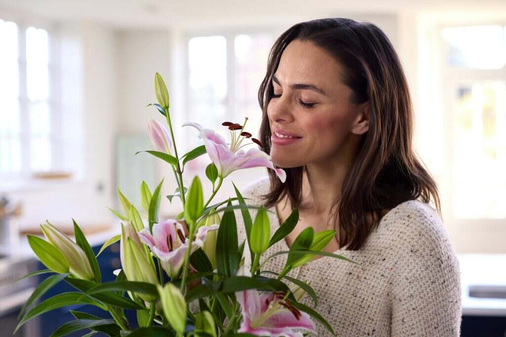 Smiling Woman At Home Smelling And Arranging Bouquet Of Flowers In Glass Vase