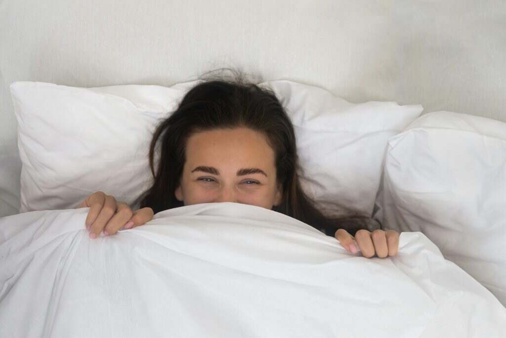 In soft cocoon of her bed, a contented woman wears a morning smile, hiding beneath a white blanket