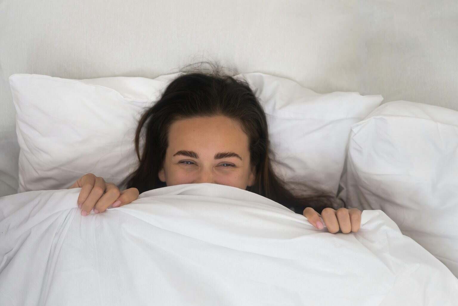 In soft cocoon of her bed, a contented woman wears a morning smile, hiding beneath a white blanket