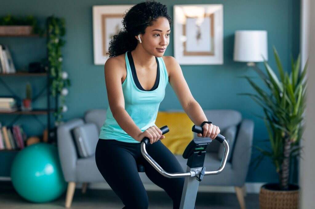 Sporty african young woman exercising on smart stationary bike and listening to music at home.