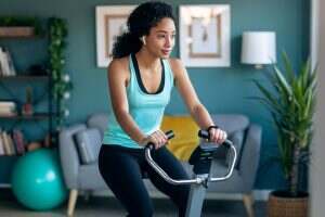 Sporty african young woman exercising on smart stationary bike and listening to music at home.