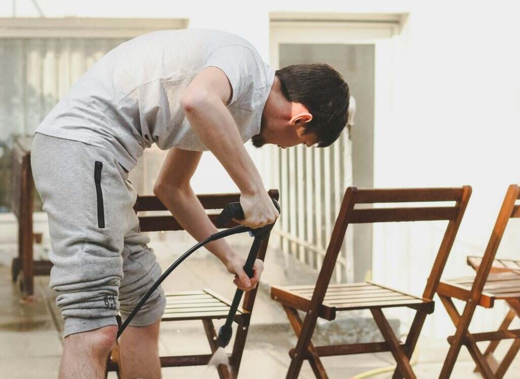 A caucasian guy washes leaning wooden garden chairs with Karcher