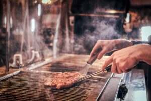 Master chef wearing uniform cooking delicious beef steak on a kitchen in a restaurant