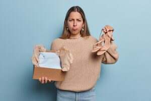Upset young European woman holds box with clothing and high heeled sandals looks unhappily at camera