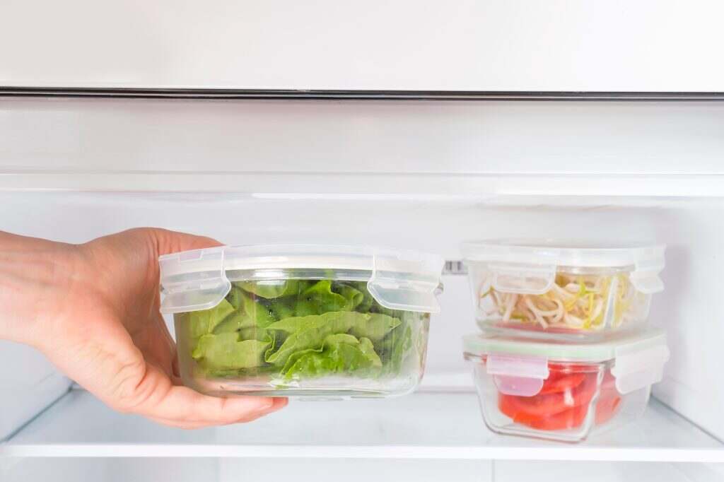 Female hand holds a glass box with green salad on the background of the freezer close-up.