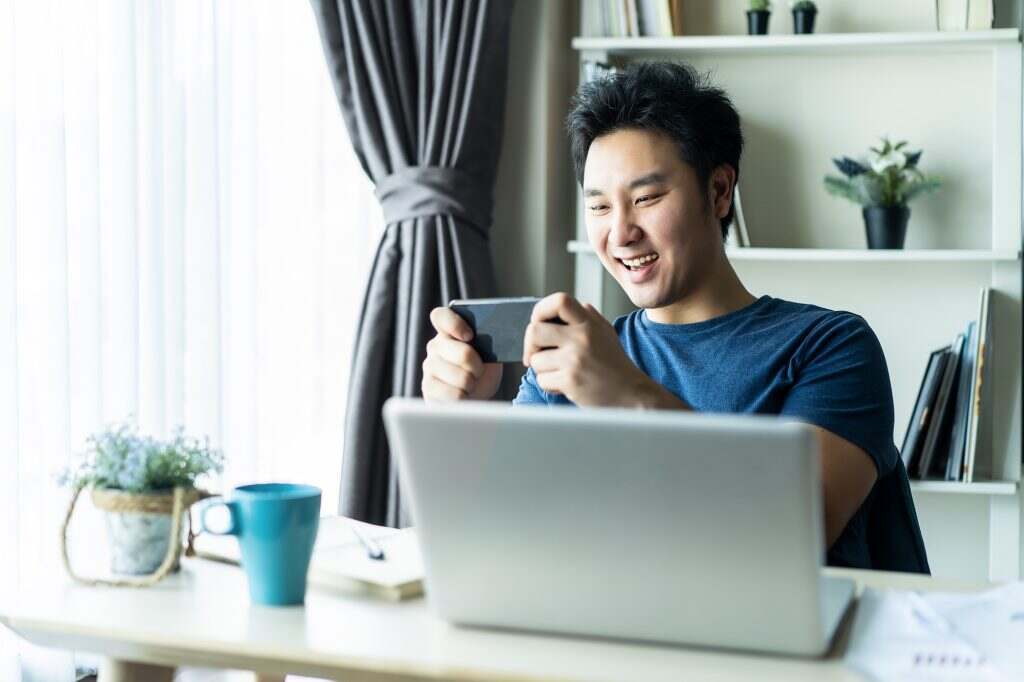 Asian young man playing online game on mobile smartphone in living room after complete work.