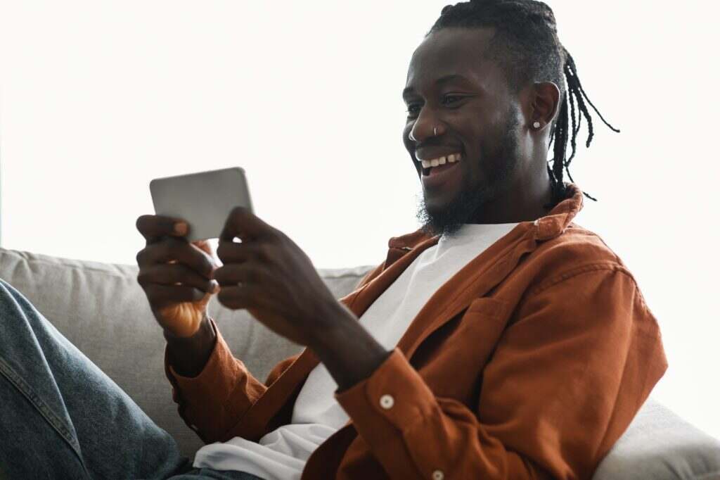 Excited african american male gamer playing games on smartphone while chilling on couch at home
