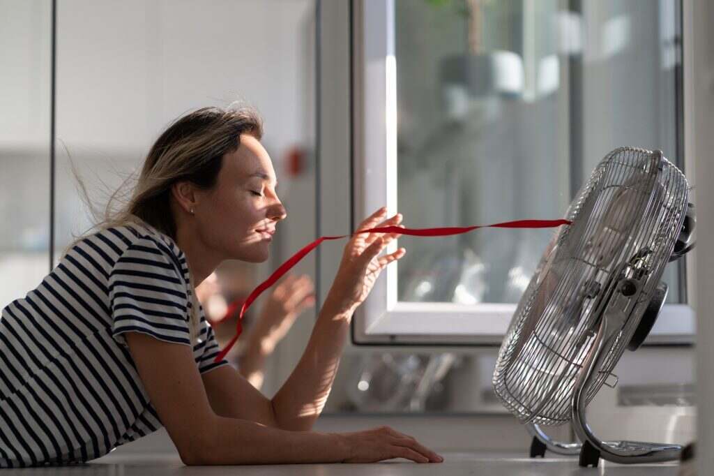 Young woman in striped t-shirt tries to catch airflow from electric fan directing to blonde hair