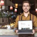 handsome young waiter holding laptop with airbnb website on screen in cafe