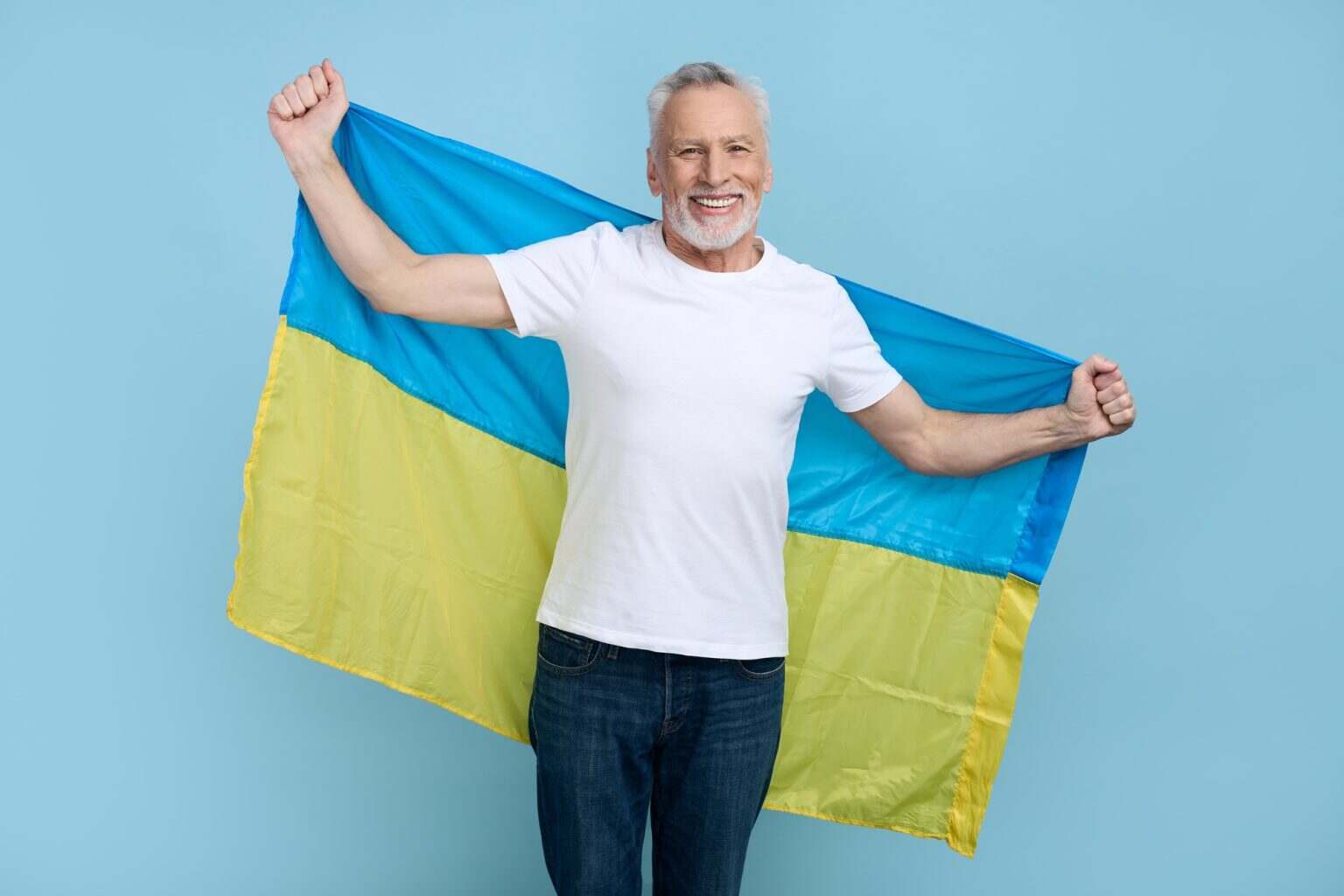 Senior man, volunteer patriotic citizen smiles at camera, carrying flag of Ukraine, isolated on blue