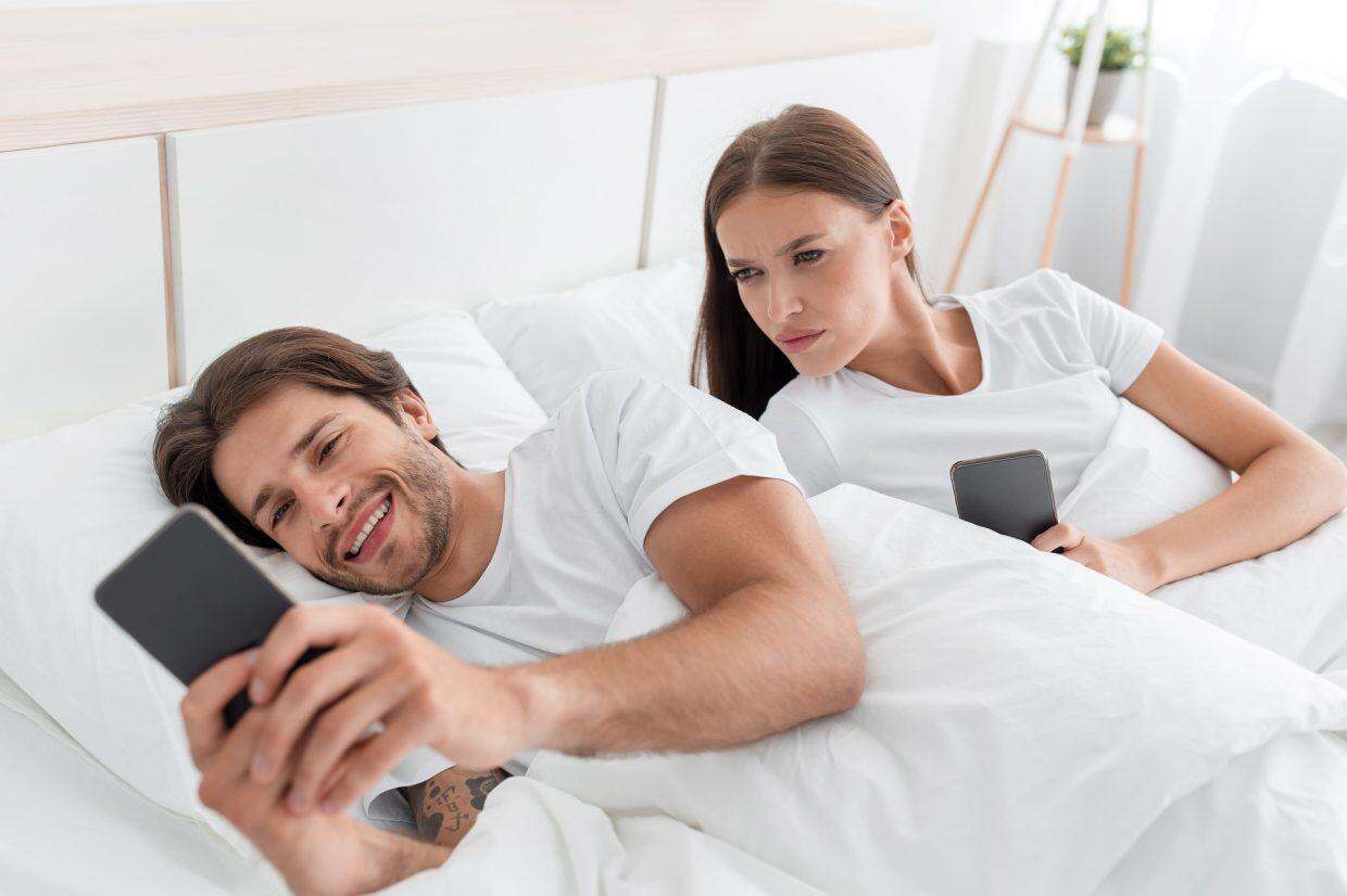 Offended angry young european wife looks at husbands phone and jealous, on bed in bedroom interior