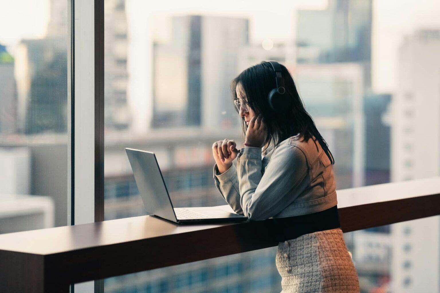 Quels profils d’hôtes et hôtesses pour votre entreprise ? Young woman with headphones using a laptop by a city window, engaged in remote work.