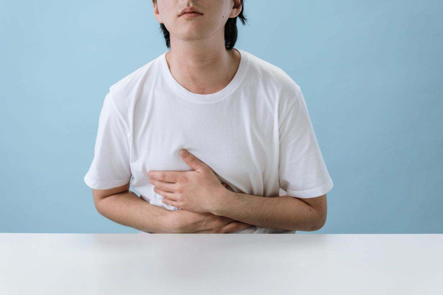 Révolution digestive : comment l’intestin transforme votre bien-être quotidien An adult man in a white shirt holds his stomach, indicating pain and discomfort, with a light blue background.
