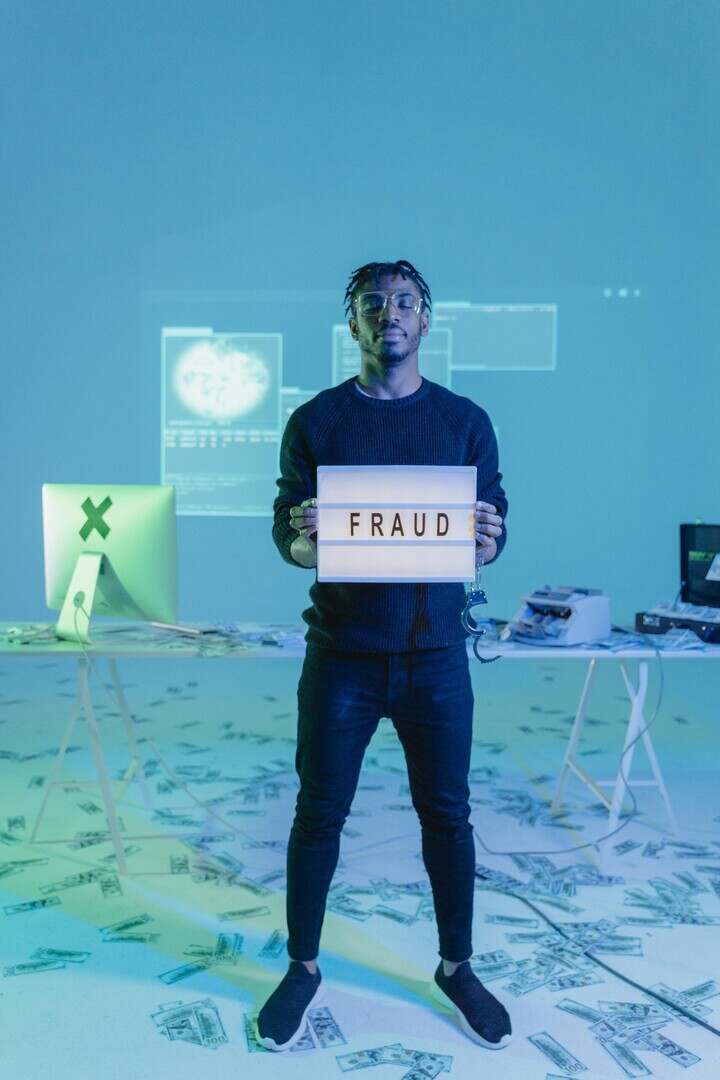 Young man holding a 'fraud' sign in a tech-focused setting with digital and cash elements.