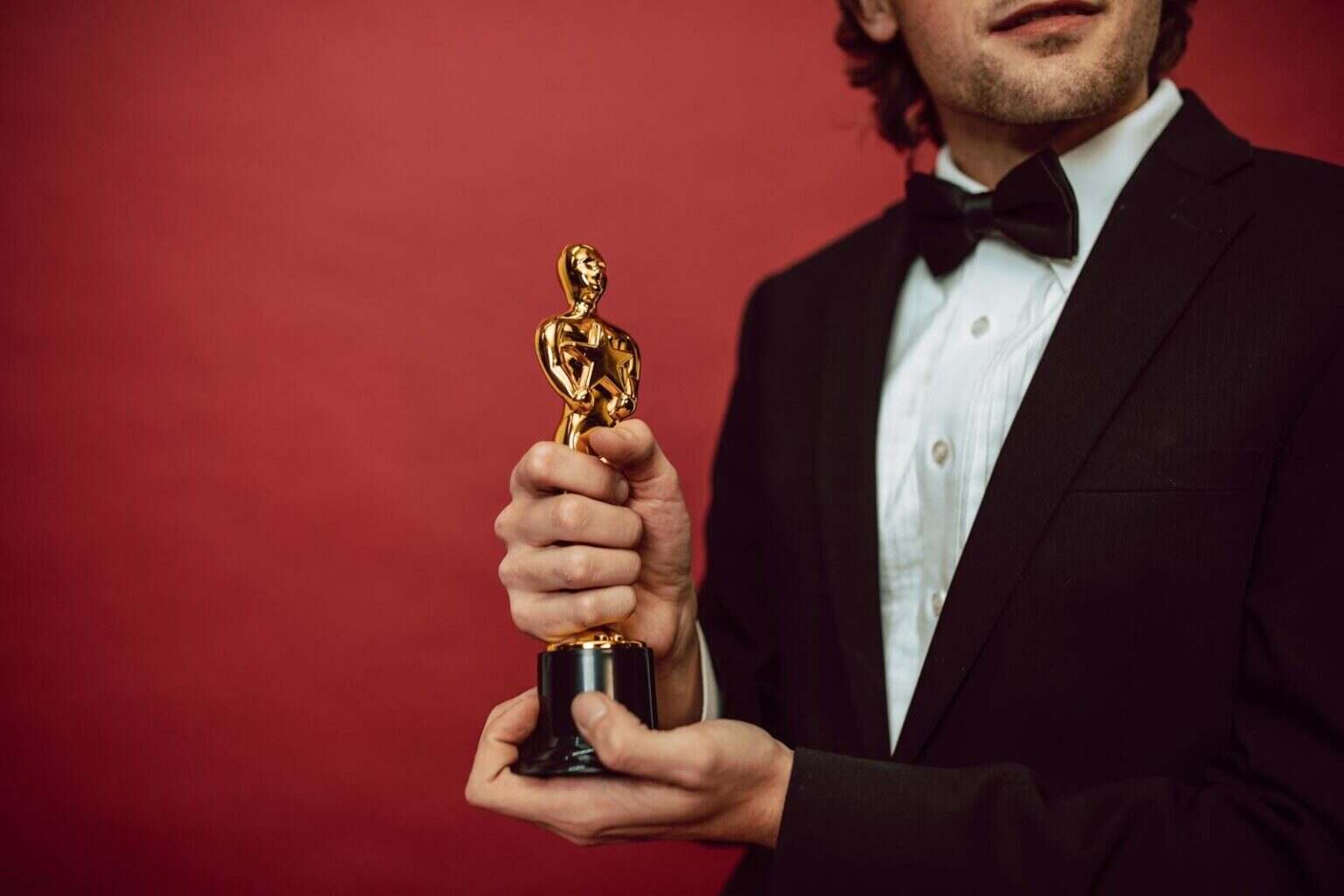 Elegant man in tuxedo holding a gold trophy against a red background, celebrating an achievement.