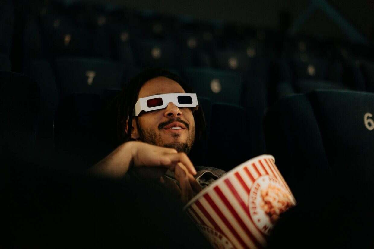 A man wearing 3D glasses enjoys popcorn while watching a movie in a cinema.