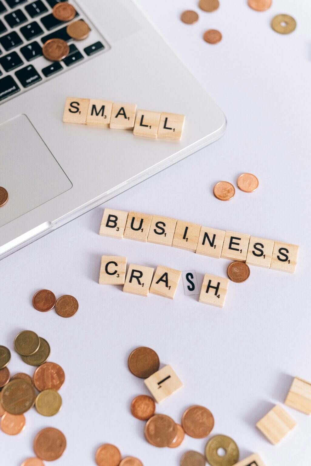 Scrabble tiles spelling 'Small Business Crash' with coins on a laptop, symbolizing financial struggle.