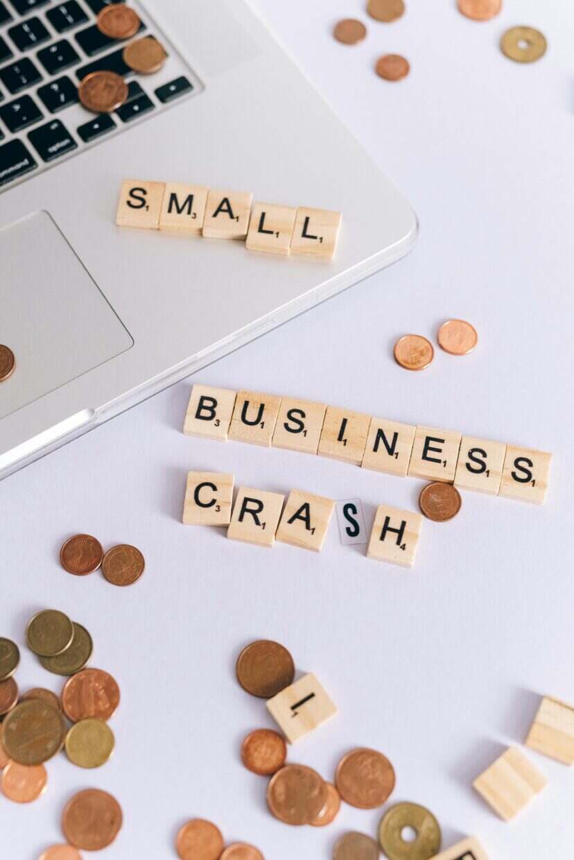 Scrabble tiles spelling 'Small Business Crash' with coins on a laptop, symbolizing financial struggle.
