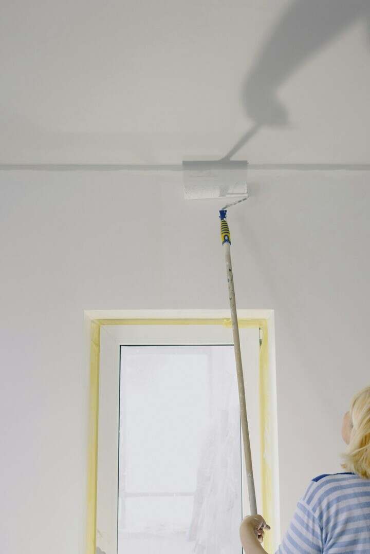A woman paints a white ceiling using a roller in a modern apartment interior.
