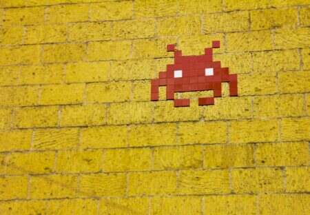 Pourquoi on ne peut pas s’empêcher de revenir aux années 2000 Street art featuring a retro pixelated space invader on a vivid yellow brick wall.