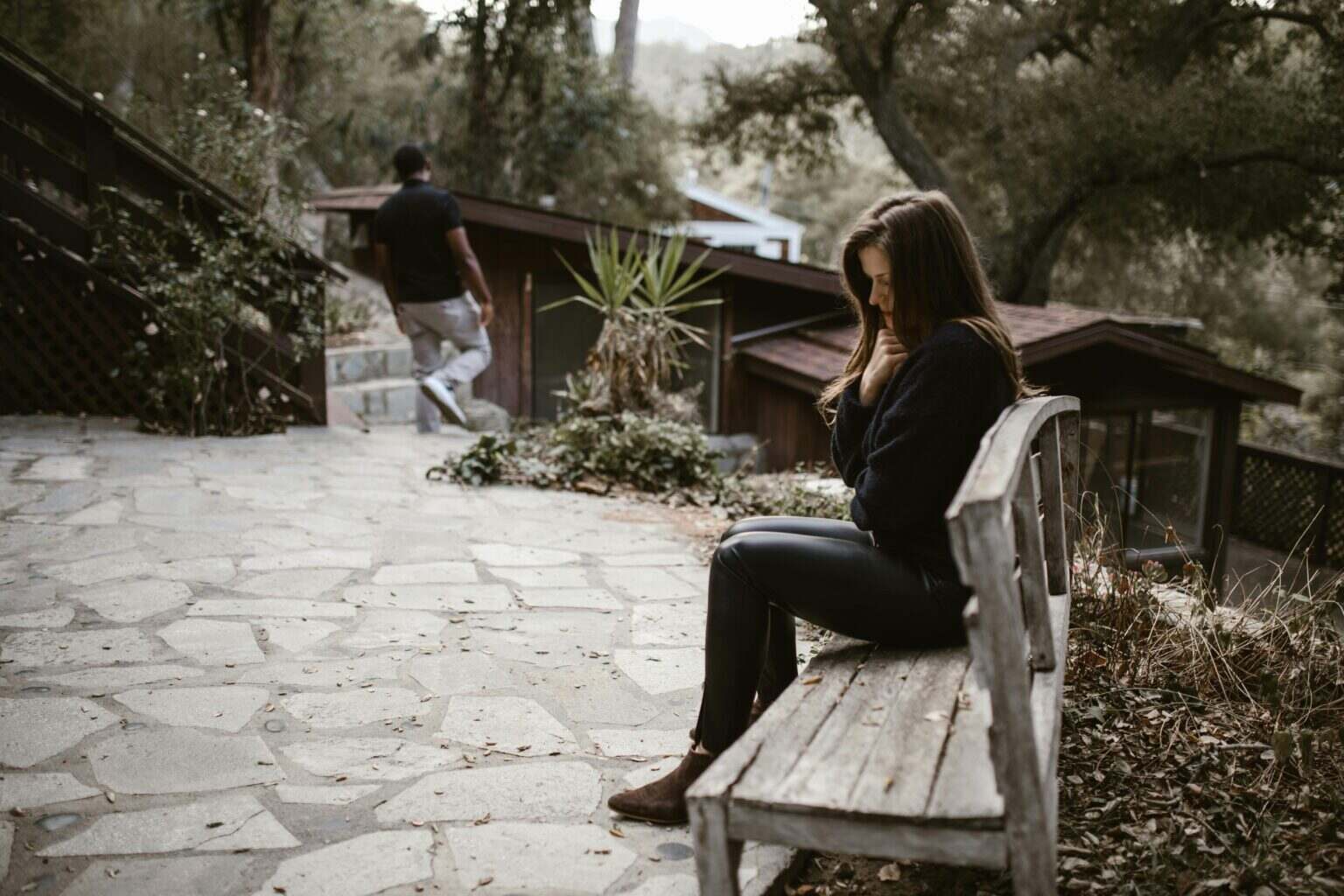 A woman sits pensively on a bench outside after a breakup, while a man walks away.