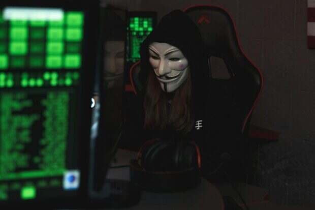 YggTorrent détruit par un hacker : la chute spectaculaire du géant français du torrent A mysterious hacker wearing a Guy Fawkes mask and black hoodie in a dimly lit room focused on computer screens.