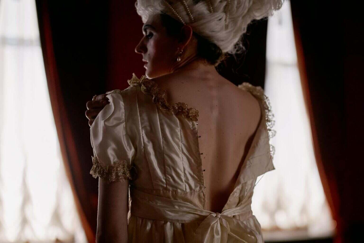A woman in historical attire stands with her back turned in a dimly lit room.