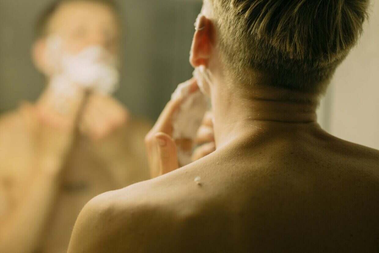 Back view of a man shaving in front of a bathroom mirror, focusing on reflection.