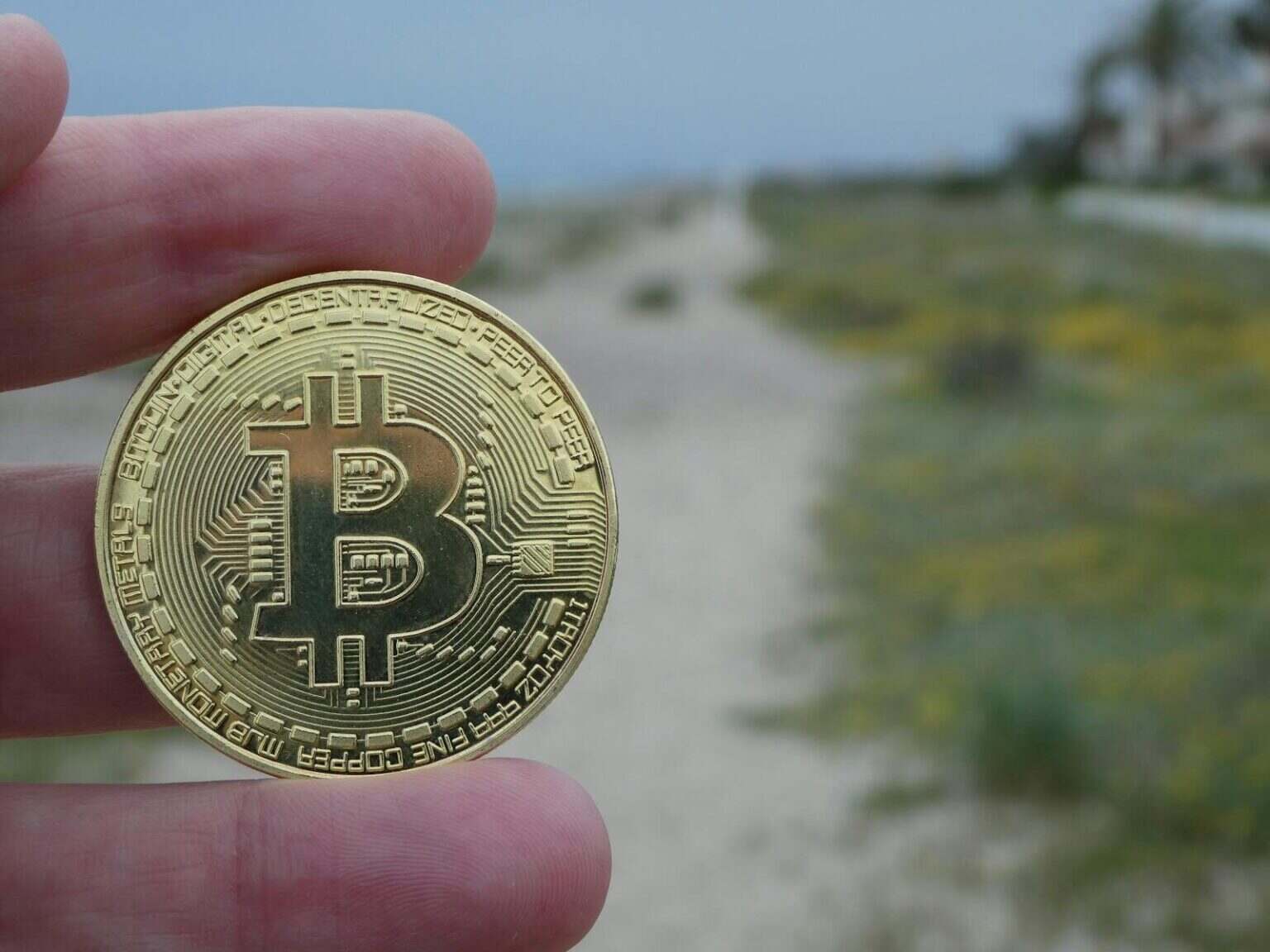 A close-up shot of a gold Bitcoin coin held by a hand with a beach background.