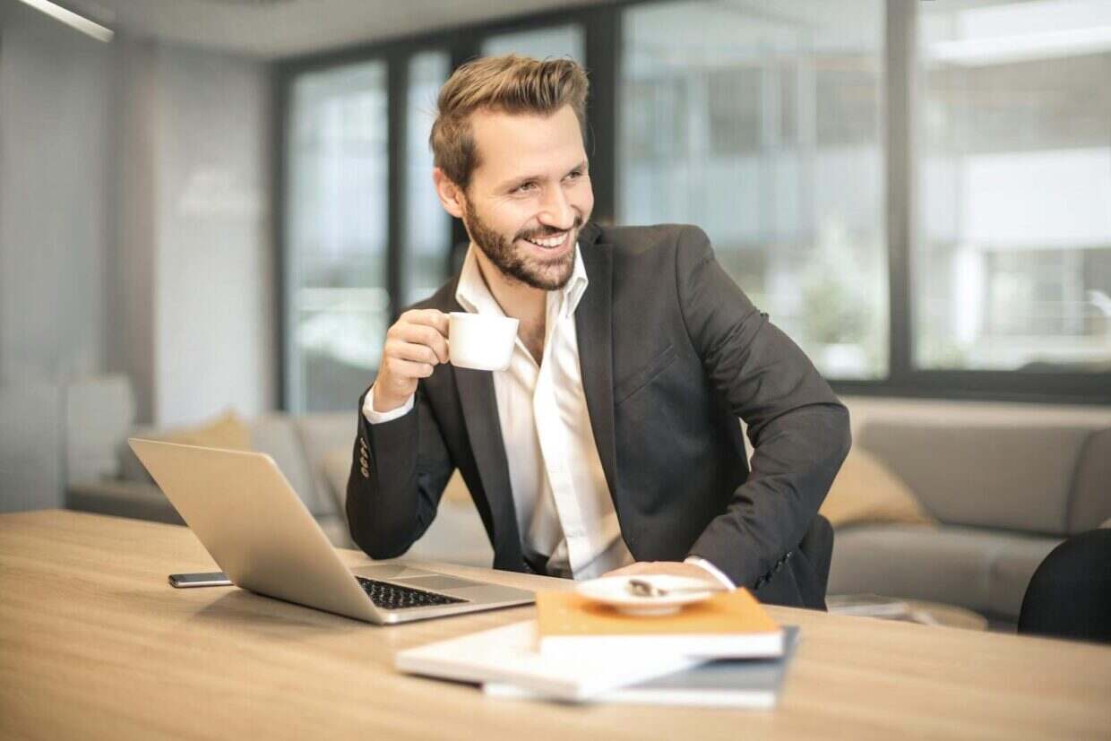 Smiling businessman in formal attire enjoying coffee break at modern office desk with laptop.