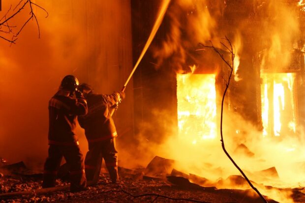 Two firefighters combat a raging fire at night, spraying water to control the flames.