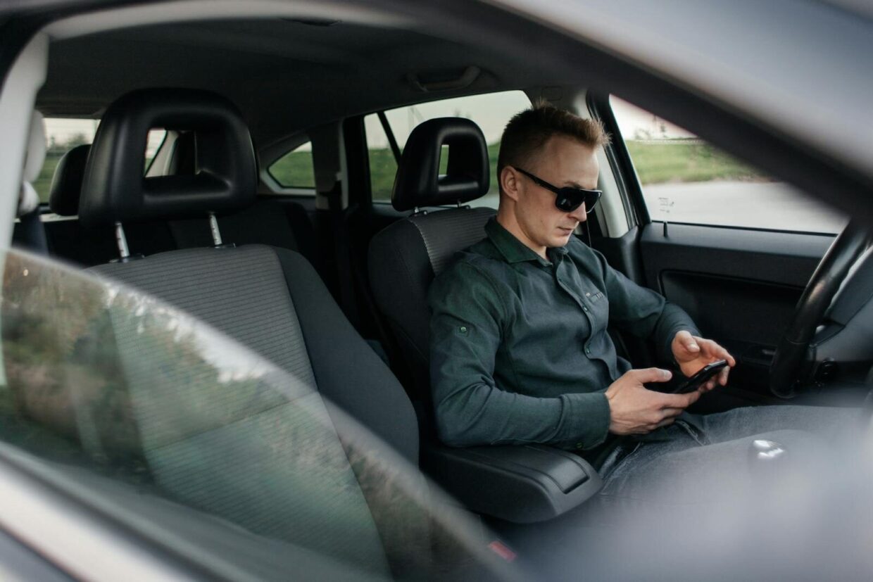 Young man using smartphone inside a parked car, wearing sunglasses.
