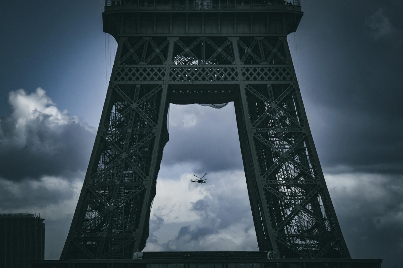 Dramatic view of a helicopter flying beneath the Eiffel Tower with a moody sky backdrop in Paris, France.