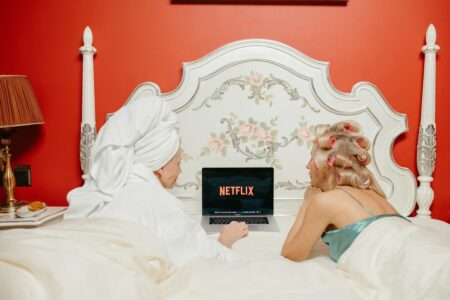 Two women lying in bed watching Netflix on a laptop, creating a cozy and bonding atmosphere.
