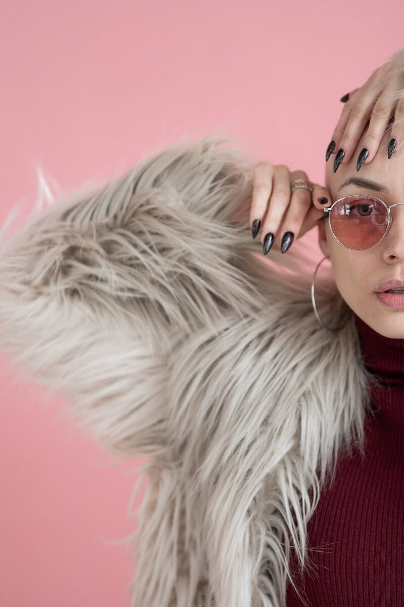 Crop stylish female with short hair wearing trendy gray fur coat looking at camera while standing on pink background with hand on head