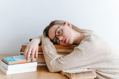 Tired female student in casual clothes lying on table with stack of books and looking away in light room in daytime