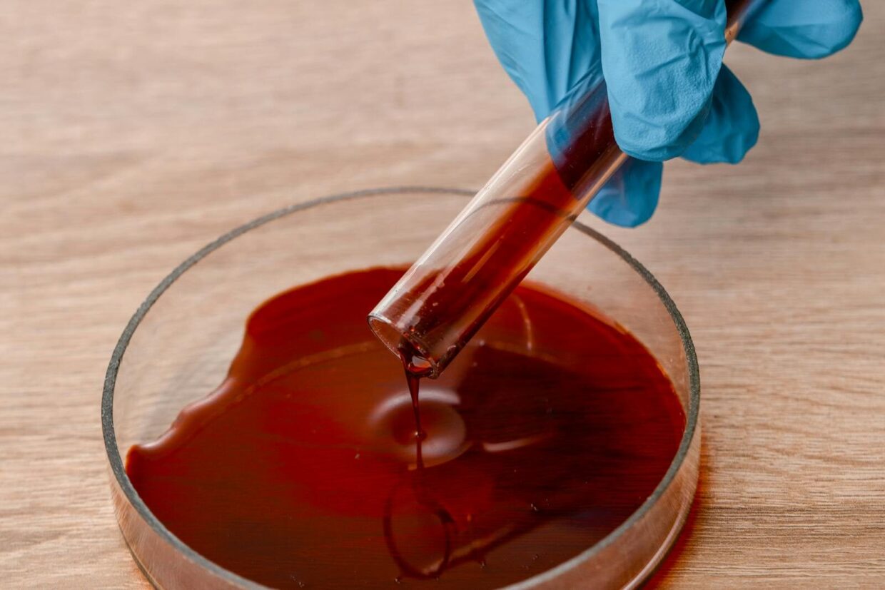 Close-up of gloved hand pouring red liquid from a test tube into a petri dish during a scientific experiment.