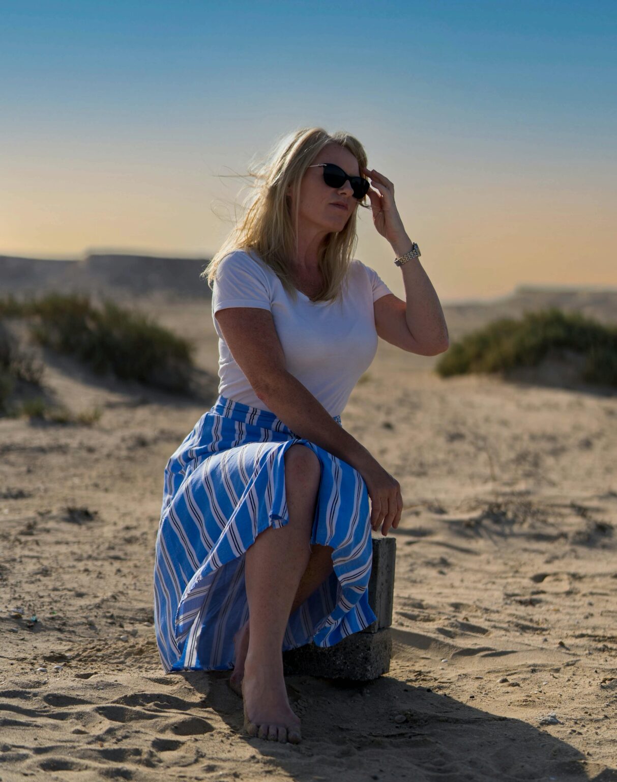 Full body of confident barefoot adult lady in summer clothes and sunglasses sitting on stone on sandy shore near grass under cloudless sky in daytime
