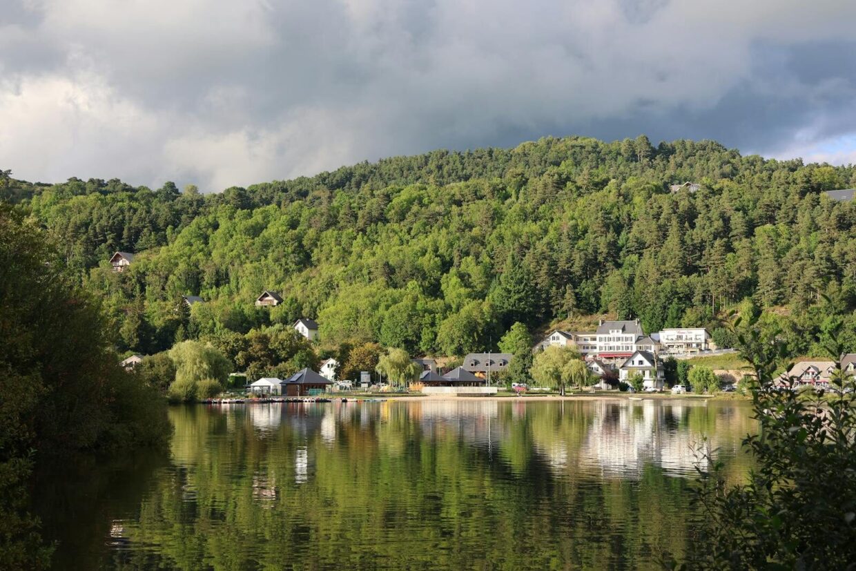 Charming village houses reflected in a serene forest-surrounded lake under a cloudy sky.