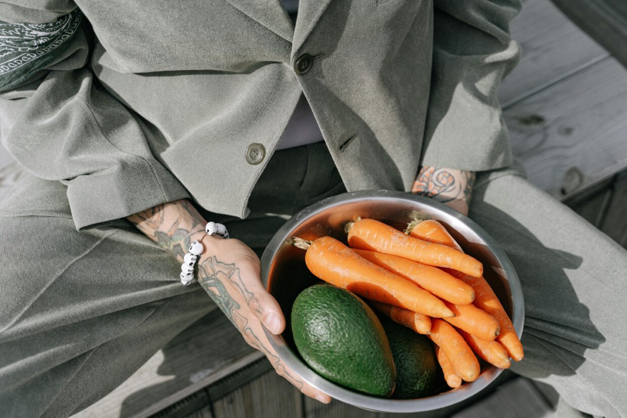 Person in gray suit holding a bowl of fresh carrots and avocados outdoors.