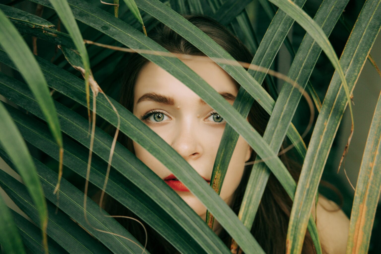 Dix marques de beauté éco-responsables pour briller cet été (sans bousiller la planète) Close-up of a woman's face partially hidden by green leaves for an artistic look.
