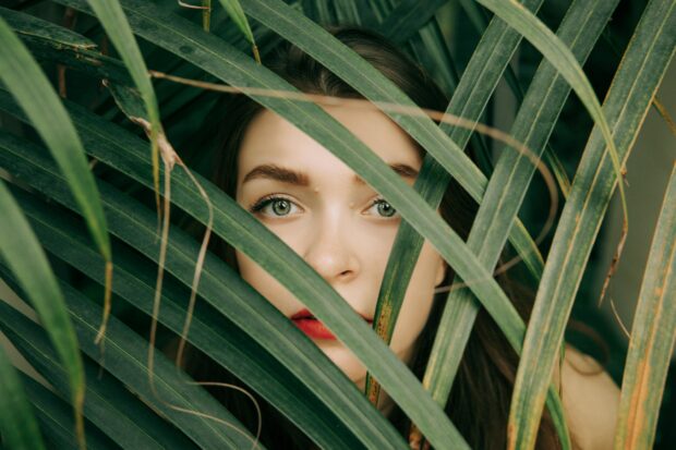 Close-up of a woman's face partially hidden by green leaves for an artistic look.